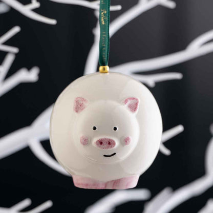 Irish Christmas | Belleek Pottery Pig Hanging Ornament suspended by a dark green ribbon with a gold connector, set against a black background with white branch silhouettes.