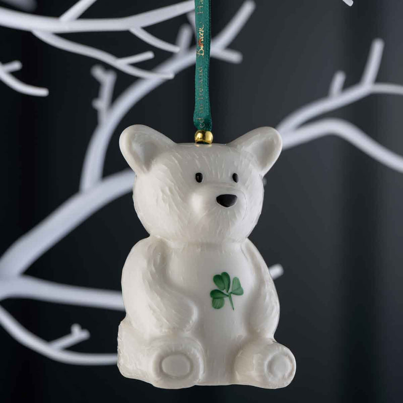 Belleek Pottery Teddy Bear Hanging Ornament with a green hanging ribbon and a small green shamrock on the chest, shown against a dark background with white branch silhouettes.