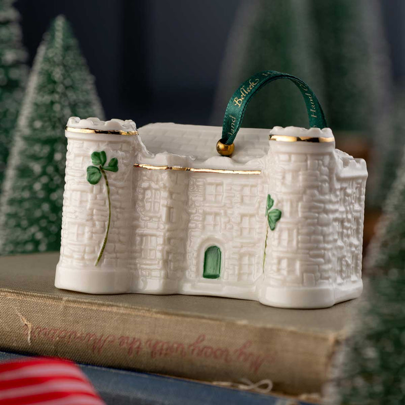 Kilkenny Castle Belleek Pottery Annual Irish Christmas Ornament 2026  with a green ribbon on a surface with Christmas trees in the background.