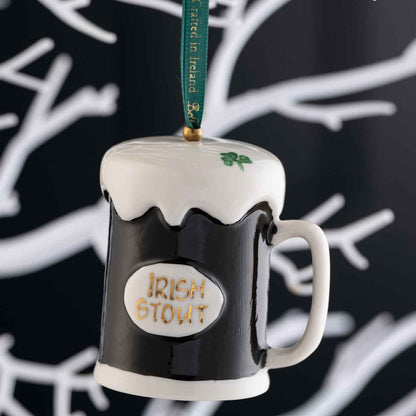 Irish Christmas Belleek Pottery Stout Hanging Ornament, and a green hanging ribbon, shown against a dark background with white branch-like patterns.