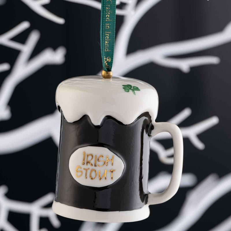 Irish Christmas Belleek Pottery Stout Hanging Ornament, and a green hanging ribbon, shown against a dark background with white branch-like patterns.