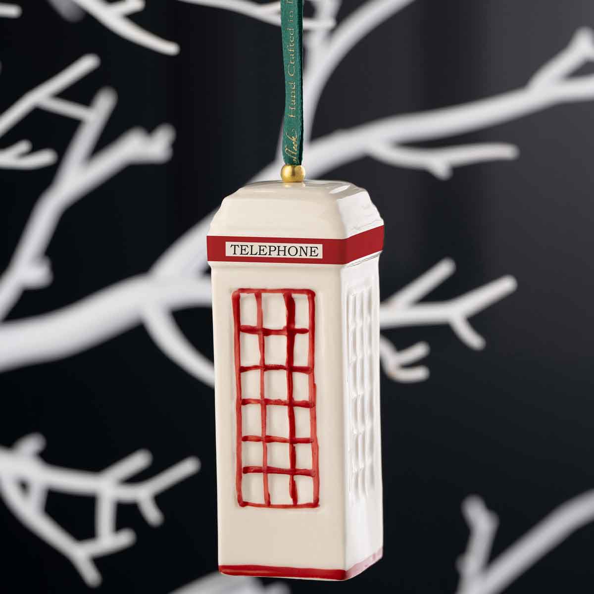 irish christmas belleek pottery traditional irish telephone box ornament with red coloring hanging from a green ribbon with a gold loop, set against a dark background with white tree branches.