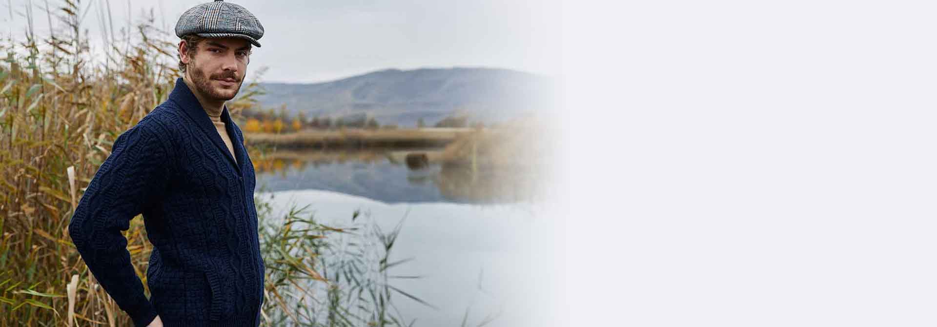 Man wearing a navy Mens Aran Knit Zipper Cardigan  and flat cap standing by a lake with mountains in the background.