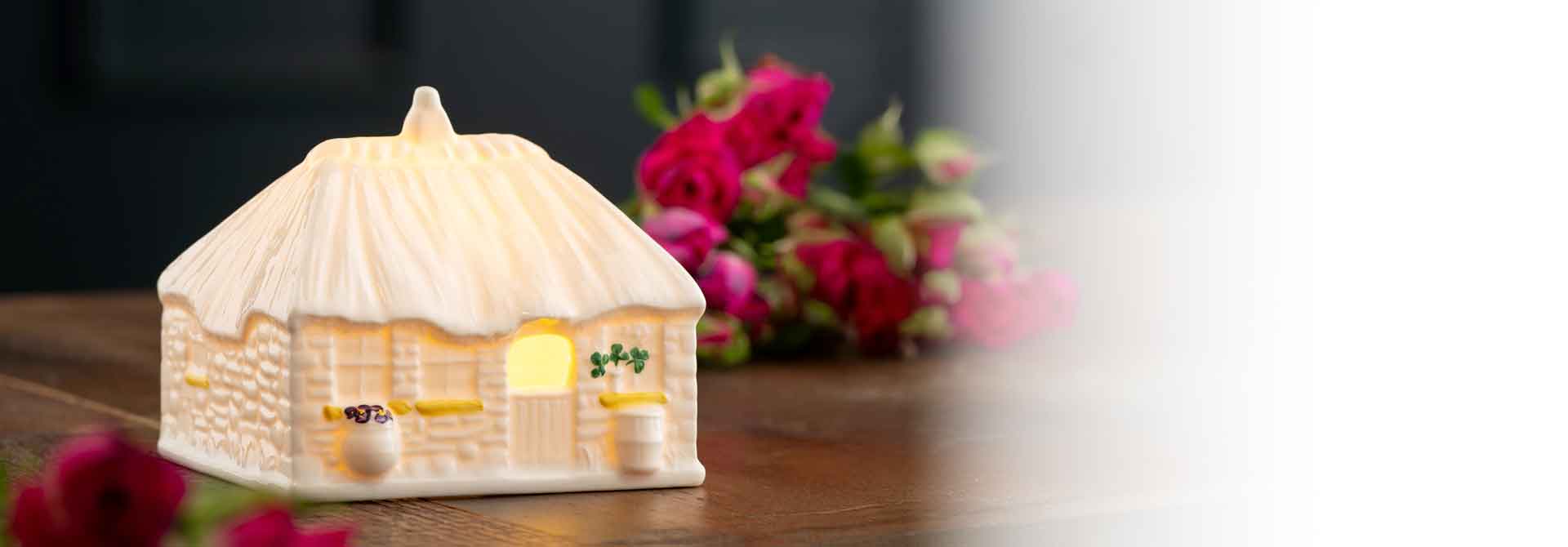A ceramic Irish cottage, illuminated from the inside, decorated with painted shamrocks and flowers on a table with roses