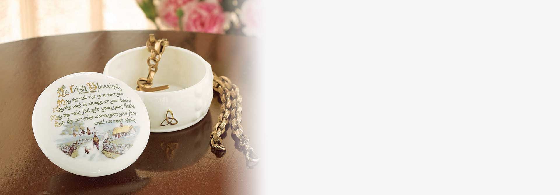 Belleek Trinket Box decorated with an Irish blessing, and gold chain on a wooden surface with floral background.