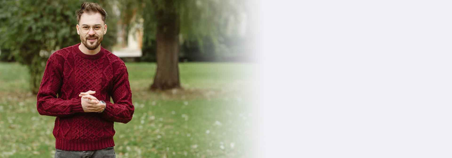 Man wearing a wine colored Irish Fisherman Cable Knit Men's Sweater standing outdoors with trees and grass in the background