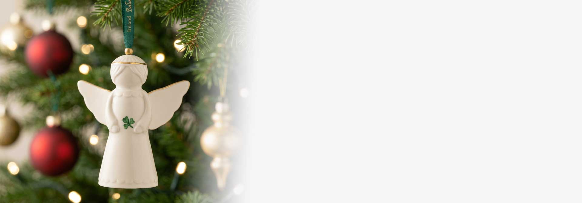 A Belleek Pottery Angel Hanging Ornament, featuring a hand painted shamrock, hanging from a green Christmas tree with red and gold ornaments and warm string lights.