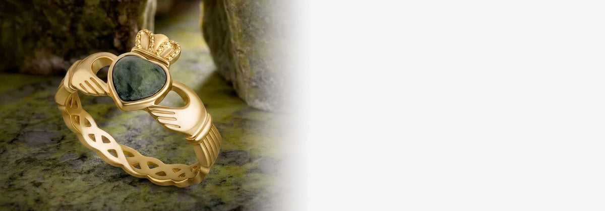 A gold Claddagh ring with a green heart-shaped stone, displayed on a textured green surface with blurred rocks in the background.