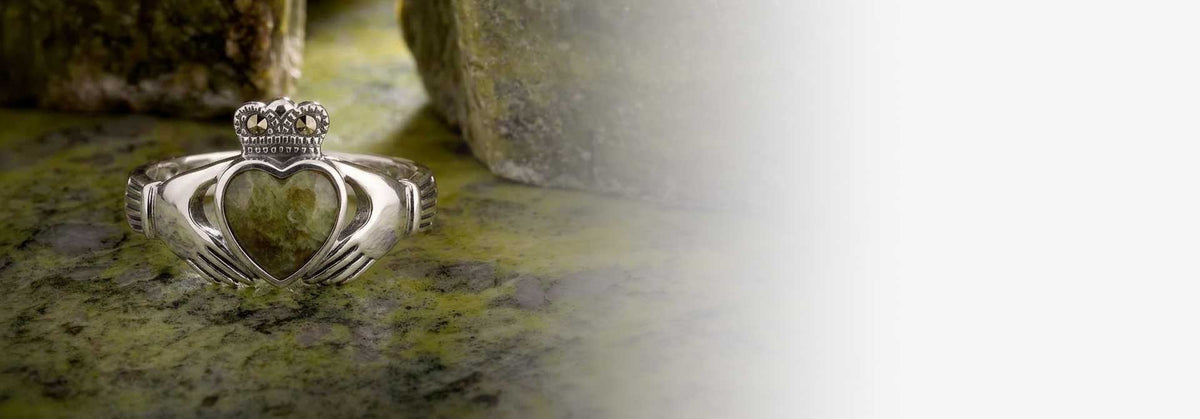 An Irish Claddagh Ring in Sterling Silver with Connemara Marble heart and marcsite stones, on a marble surface with a soft-focus blurred background.