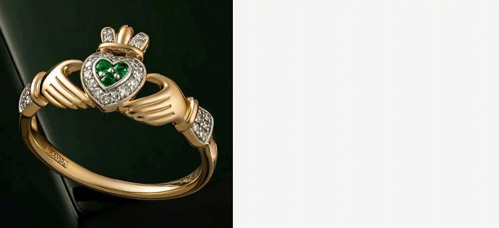 A gold Claddagh ring with a heart of Emeralds surrounded by Diamonds on a dark green surface