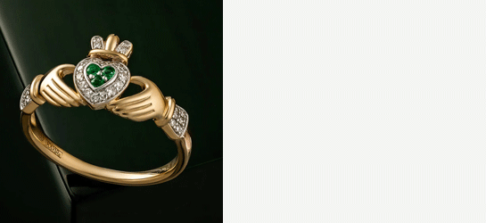 A gold Claddagh ring with a heart of Emeralds surrounded by Diamonds on a dark green surface