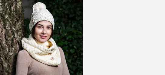 Woman wearing an Aran Knit hat & scarf in color natural, standing against a tree