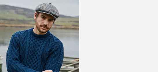 Man wearing a blue Aran knit sweater and Irish tweed cap standing in front a lake & mountains