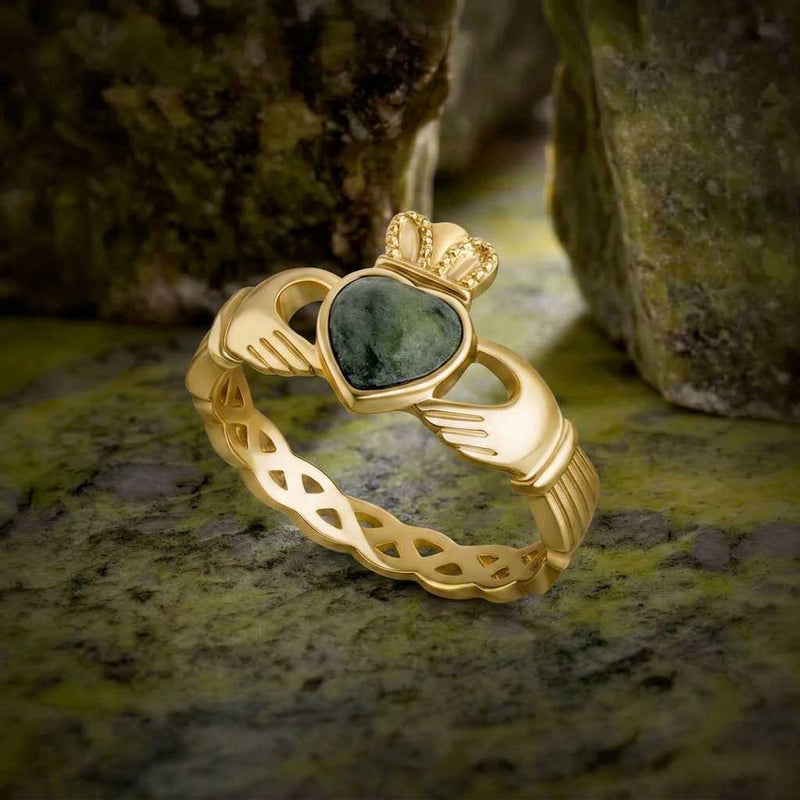 A 10K Gold Connemara Marble Celtic Band Irish Claddagh Ring with Celtic Knot band, displayed on a marble with blurred rocks in the background.