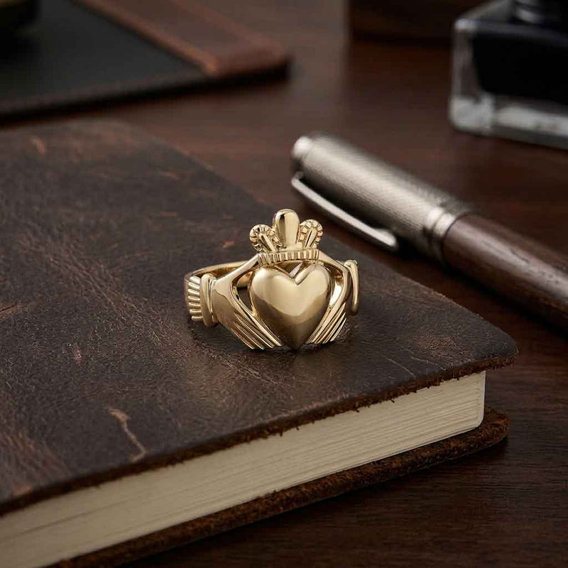 An Irish Large 14k Gold Mens Claddagh Ring, resting on a dark brown leather notebook next to a silver pen on a wooden desk.
