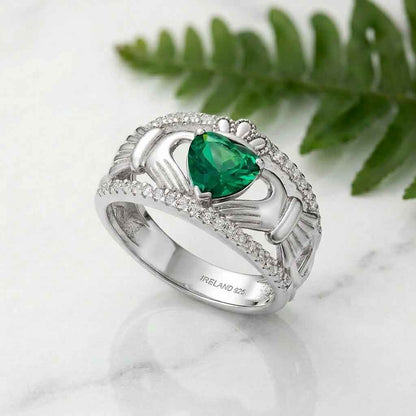 Sterling Silver Celtic Band with Green Crystal Claddagh Ring on a marble surface with fern leaves in the background