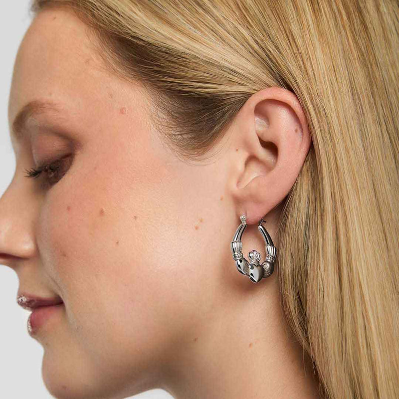 Close-up of a woman wearing Sterling Silver Medium Hoop Claddagh Earrings on a white background