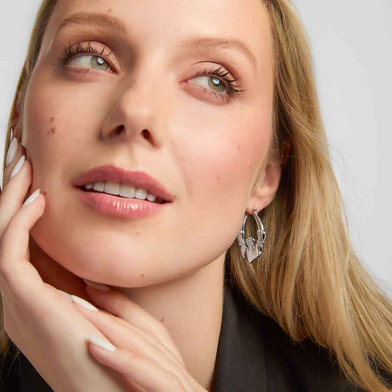 Close-up of a woman wearing Sterling Silver Large Hoop Claddagh Earrings 
 with a neutral background