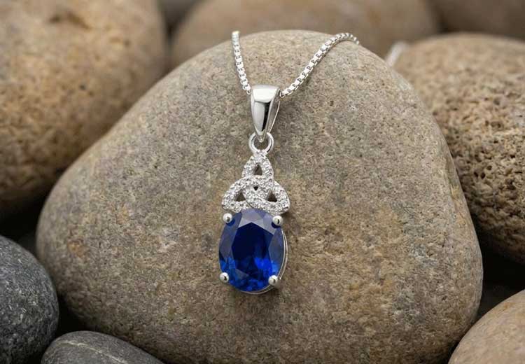 A Celtic Trinity Knot Birthstone Necklace with Blue Sapphire Crystal Pendant resting on smooth brown rocks