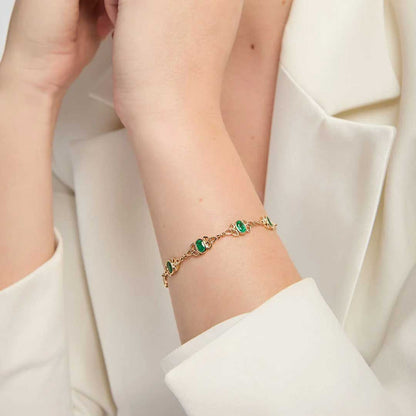 An Irish 14k Gold Emerald Trinity Knot Bracelet, worn on a woman's wrist. The image is cropped to show the person's hands and arms resting in a white blazer.
