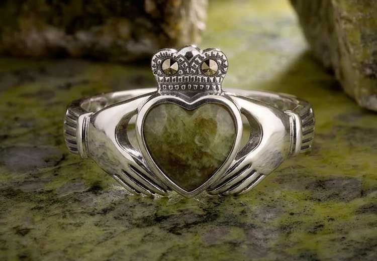 An Irish Claddagh Ring in Sterling Silver with Connemara Marble heart and marcsite stones, on a marble surface with a soft-focus blurred background.