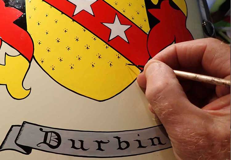 Hand painting a coat of arms gift with the 'Durbin' arms and name, using a paintbrush.
